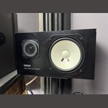 Černý studiový monitor s dřevěným povrchem. Na přední straně je bílý basový reproduktor a menší výšek s kovovou mřížkou. V levém dolním rohu je bílý…