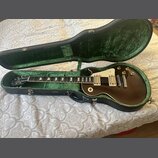 Tmavně hnědá elektrická kytara typu Les Paul s krémovým pickguardem a dvěma humbuckery v černém tvrdém kufru s zeleným polstrováním. Na hmatníku jsou…
