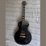 Černá elektrická kytara s gold hardwarem a bílými blokovými vložkami na hmatníku. Na hlavě je nápis Epiphone a logo, na snímači je vidět nápis Epipho…