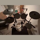 Elektronická bicí souprava s černými pady a činely na kovovém stojanu. Na bílém snare padu je vidět nápis Roland. Soubor obsahuje pět padů a dva čine…