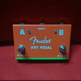 Oranžový efekt pedál s nápisem Fender a označením ABY PEDAL. Na horní části jsou dva vstupy/výstupy označené písmeny A a B a nápis IN/OUT. V dolní čá…