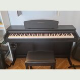 Černý digitální piano v černém dřevěném korpusu s přívodní stoličkou v odpovídající barvě. Na horní části krytu i v pravém dolním rohu je bílý nápis…