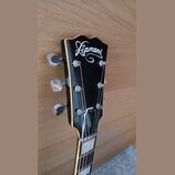 Detail hlavy černé kytary s bílým nápisem Diamant. Na hlavě jsou vidět tři chromované ladičky a struny vedené k maticím saddles. Povrch je glossy, s…