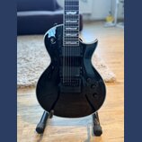 Černá elektrická kytara s lesklým povrchem a bílým lemováním korpusu. Na snímači je viditelný nápis EMG. Kytara má dva humbuckery, tři ovládací knofí…