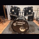 Černá bicí souprava tvořená dvěma tommy a basovým bubnem. Na basovém bubnu jsou viditelné nápisy SONOR, SHURE, METALO a bern, dále logo National Drum…