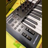 Černý MIDI kontrolér s klávesnicí. Na šasi jsou viditelná loga M-AUDIO a OXYGEN25. Nástroj obsahuje několik otočných potenciometrů, tlačítka s nápisy…