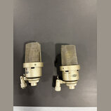 Dva kondenzátorové mikrofony stříbrné barvy s kovovými mřížkami. Na těle pravého mikrofonu je nápis -10dB 0 20dB a logo NEUMANN. Levý mikrofon má na…