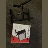 Černý kovový stojan na zesilovač s logem Fender na jedné z nožních gumových konцоvek. Vedle stojanu je umístěna originální krabice s nápisem Fender A…
