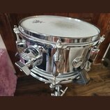 Stříbrný kovový snare drum s bílými koženem. Na korpusu je viditelný oválný štítek s nápisy MAPEX, MPX a STEEL. Nástroj vykazuje mírné znečištění pov…