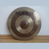 Kovový činel zlatavé barvy s viditelným Oxidováním a tmavými skvrnami v prostřední části. V horní části je černý nápis istanbul Agop Handmade cymbals…