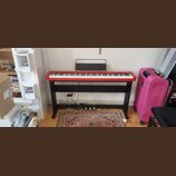 Digitální piano s červeným šasi a černými klávesami, umístěné na černém dřevěném stojanu. Nástroj má přiložený černý notový pult. Na povrchu nástroje…