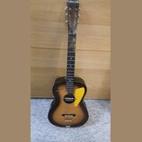 Akustická kytara s tmavým, přechodovým povrchem (sunburst). Na hlavě je bílý nápis CABALLERO a na korpusu vlevo od otvoru nápis astano. Nástroj má žl…