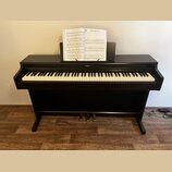 Černý digitální piano s bílými a černými klávesami. Nad klaviaturou je umístěno logo YAMAHA a na notovém pultu jsou položeny notové listy. Nástroj st…