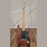 Elektrická kytara s hnědým dřevěným korpusem a světle hnědým javorovým hmatníkem. Na hlavě je viditelné logo Fender. Kytara má černý mramorovaný pick…