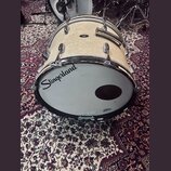 Bílá basová bubna s kovovým kováním a madlem. Na přední blaně je černý nápis Slingerland a malé logo Remo. Povrch korpusu vykazuje známky opotřebení…