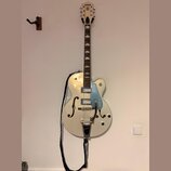 Bílá polodupatá elektrická kytara s modrým pickguardem a chromovaným hardwarem. Na hlavě hmatníku je nápis GRETSCH a na pickguardu je svisle umístěný…
