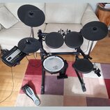 Elektronická bicí souprava s černým rámem a černými pady. Na modulu, na snare padu i na pedálu je vidět bílý nápis Roland. Souprava obsahuje pět padů…