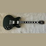 Černá polodupatá elektrická kytara s bílým lemováním a hnědými f-otvory. Na hlavě je viditelné logo Gibson. Nástroj má dva humbuckery, tři ovládací k…
