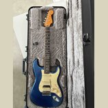 Modrá elektrická kytara typu Stratocaster s bílým pickguardem a javorovým hmatníkem v otevřeném černém tvrdém kufru s šedým polstrováním. Na hlavě ná…