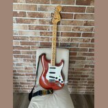 Elektrická kytara typu Stratocaster v červeně hnědé barvě s bílým pickguardem a světle hnědým hnedobým hmatníkem. Na hlavě je viditelné logo Fender S…