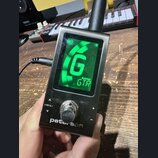 Kompaktní digitální ladič stříbrné barvy s displejem. Na spodní části je bílý nápis peterson a nad ním ovládací prvky MENU, +, - a kovové kolečko. Na…