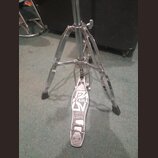 Chromovaný kovový stojan s pedálem, pravděpodobně hi-hat. Na pedálu je viditelné logo TAMA a nápis THE LEGEND IN INNOVATION. Povrch pedálu vykazuje z…