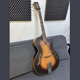 Kytara typu archtop s tmavě hnědým sunburst povrchem a bílým lemováním. Na hlavě jsou vidět čtyři ladičky a dva červené dekorativní prvky. Tělo má dv…