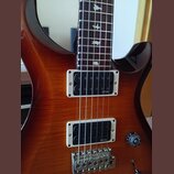 Elektrická kytara s hnědým lakovaným povrchem s viditelnou strukturou dřeva. Nástroj má dva snímače typu humbucker s bílými rámečky, na kterých je uv…