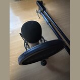 Kondenzátorový mikrofon s černou pěnovou windscreen v černém pavoucím držáku. Mikrofon je umístěn před kulatým černým popfiltrem. Vedle sestavy leží…