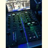Černý DJ kontrolér s několika knoflíky, posuvníky a podsvícenými tlačítky. Na přístroji je svisle napsán bílý text TRAKTOR Z1. V pozadí je vidět zapn…