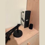 Na fotografii je krabice od mikrofonu s nápisy SHURE, MV7 a MV7 PODCAST MICROPHONE. Vedle krabice je černý pěnový popfilter a černý mikrofonní ramení…