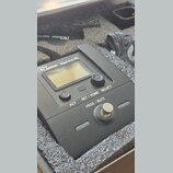 Černý elektronický přístroj s displejem, umístěný v polstrováním vystlaném pouzdře. Na horní hraně je nápis the ut.bone GigA Pro PR. Zařízení obsahuj…