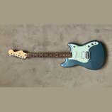Modře šedá elektrická kytara s bílým pickguardem a hnědým hmatníkem. Na hlavě je viditelné logo "Fender". Nástroj má konfiguraci snímačů dva jednoduc…