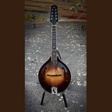 Mandolina s hruškovitým korpusem v tmavě hnědém sunburst provedení. Na hlavě je bílý nápis „12“. Nástroj stojí na černém stojanu. Povrch laku vykazuj…