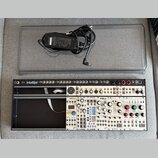 Fotografie zobrazuje Eurorack modulární syntetizátor v černém racku s bílým předním panelem a přiložený napájecí adaptér v šedém přepravním kufru. Na…