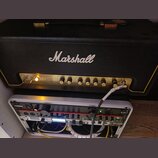 Černý kytarový kombozesilovač značky Marshall s nápisem ORIGIN 50 na zlatém předním panelu. Pod ním jsou umístěna dvě racková zařízení, z nicha horní…