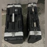 Dvě černé obdélníkové transportní skříně z texturovaného materiálu s hliníkovými hranami a zapínacími zámky. Na levé skříni je vidět čtvercové logo s…