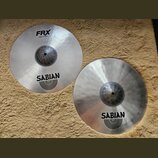 Dvě stříbrné činely značky Sabian s matným povrchem. Levý činel nese nápisy FRX, Hats 14"/35 cm a logo SABIAN. Pravý činel má pouze logo SABIAN. Oba…