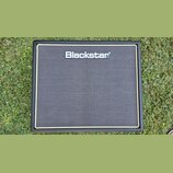 Kytarový kombozesilovač s černým povrchem a šedou textilní maskou. V horní části je umístěno stříbrné logo s nápisem Blackstar. Celý přístroj je obal…