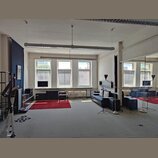 Interiér místnosti s šedým kobercem, bílými stěnami a velkým zrcadlem na pravé straně. V prostoru je umístěna černá kožená pohovka, červený koberec a…