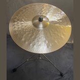 Zlatý kovový činel umístěný na černém stativu. Na povrchu je nápis „Paiste“ a označení „Dry Ride 20"“. Povrch činelu je zdobný koncentrickými rýhami…
