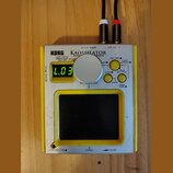 Stříbro-žlutý přenosný syntetizátor s nápisy KORG a KAOSSILATOR DYNAMIC SYNTHESIZER. Zařízení má malý digitální displej s nápisem L.03, jeden velký s…