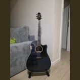 Tmavě modrá akustická kytara s cutawayem, umístěná na černém stojanu. Na hlavě hmatníku je bílé logo Takamine. Kytara má tmavý hmatník s ozdobnými vl…