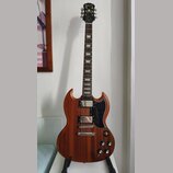 Elektrická kytara s hnědým dřevěným povrchem a černým pickguardem. Na hlavě hmatníku je bílý nápis Epiphone. Nástroj má dva humbuckery, Tune-o-matic…