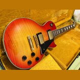 Elektrická kytara s trupem ve tvaru single-cut a povrchem v červeno-žlutém přechodu (sunburst) s viditnou strukturou dřeva. Nástroj má dva humbuckery…