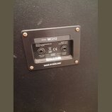 Fotografie zachycuje zadní panel reproduktoru s černým povrchem. Na stříbrném štítku je uveden nápis MODEL: MC212, made by Marshall Amplification plc…
