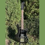 Černá elektrická kytara s černým pickguardem a hnědým hmatníkem. Na hlavě krku je viditelné bílé logo Fender. Nástroj má dva snímače, tři ovládací kn…
