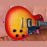 Elektrická kytara s tmelem v červeno-žlutém přechodu (sunburst) a viditnou strukturou dřeva. Nástroj má dva humbuckery s černými rámečky, černý plech…