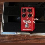 Červený efekt pedál s nápisy HALL OF FAME 2 a tc electronic. Na horní části jsou čtyři ovládací knoflíky s popisky DECAY, PRE-DELAY, TONE a LEVEL. V…