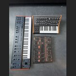 Fotografie zobrazuje tři elektronické hudební přístroje. Vlevo je šedý syntetizátor s klaviaturou a nápisem UMIKKO v levém dolním rohu. Vpravo nahoře…